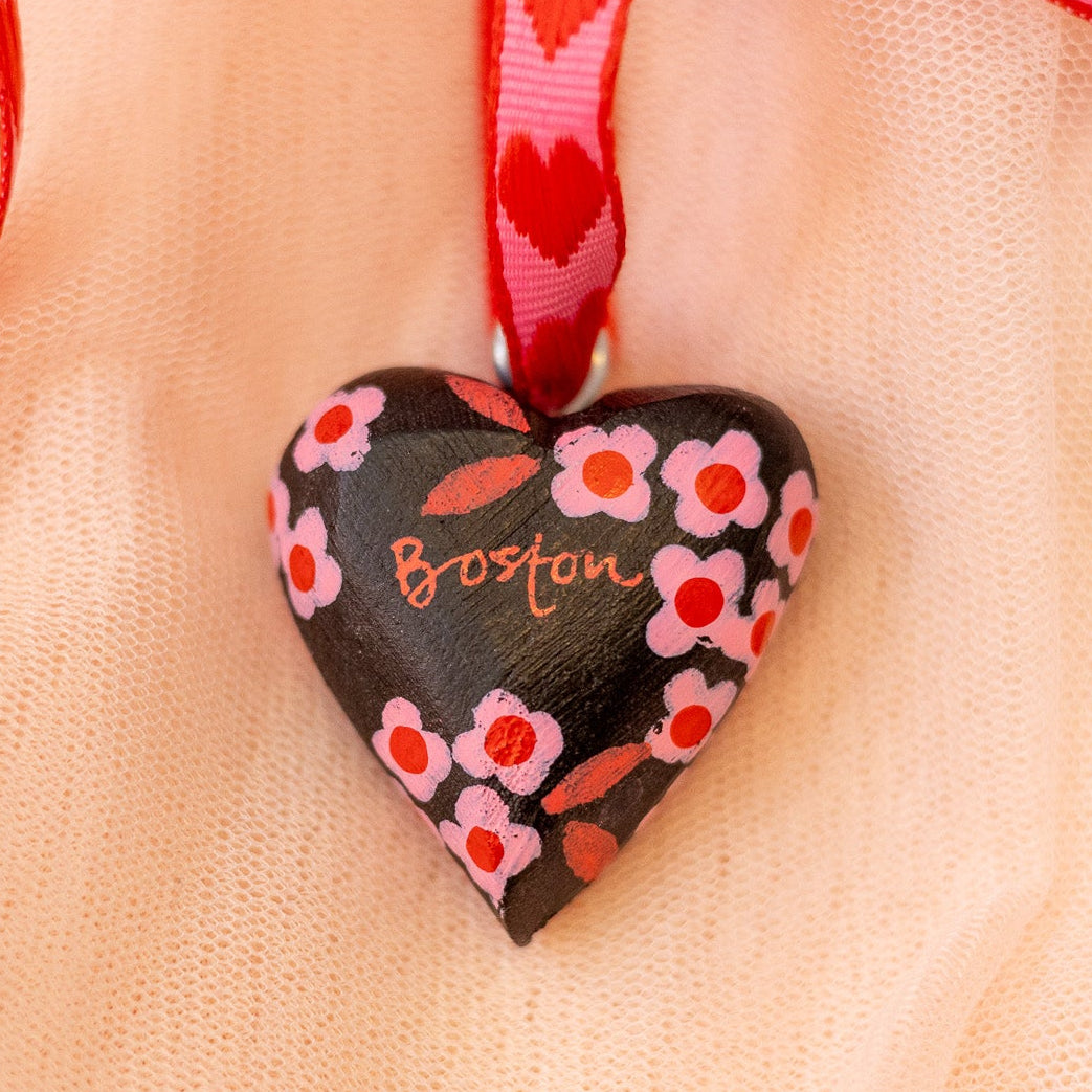 Heart-shaped pendant with floral design and 'Boston' text on a red ribbon against a beige background