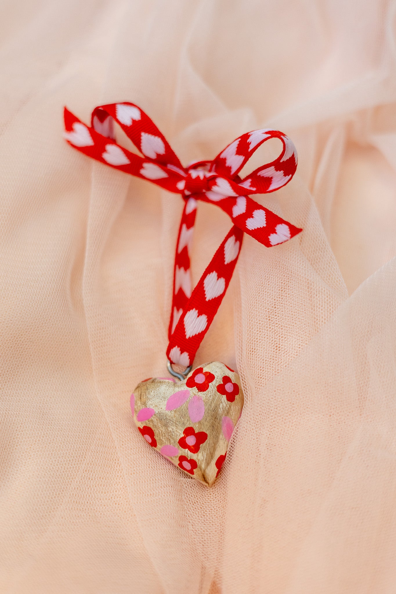 Heart-shaped wood ornament with red floral patterns and a red ribbon on a sheer peach fabric background