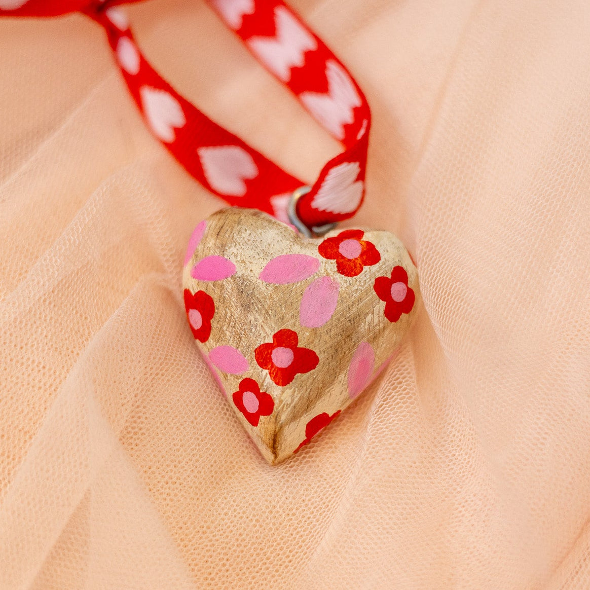 Heart-shaped wood ornament with red floral patterns and a red ribbon on a sheer peach fabric background