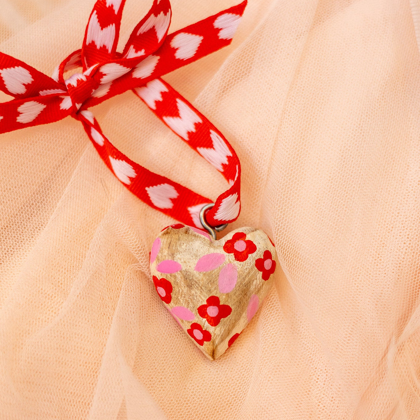 Heart-shaped wood ornament with red floral patterns and a red ribbon on a sheer peach fabric background