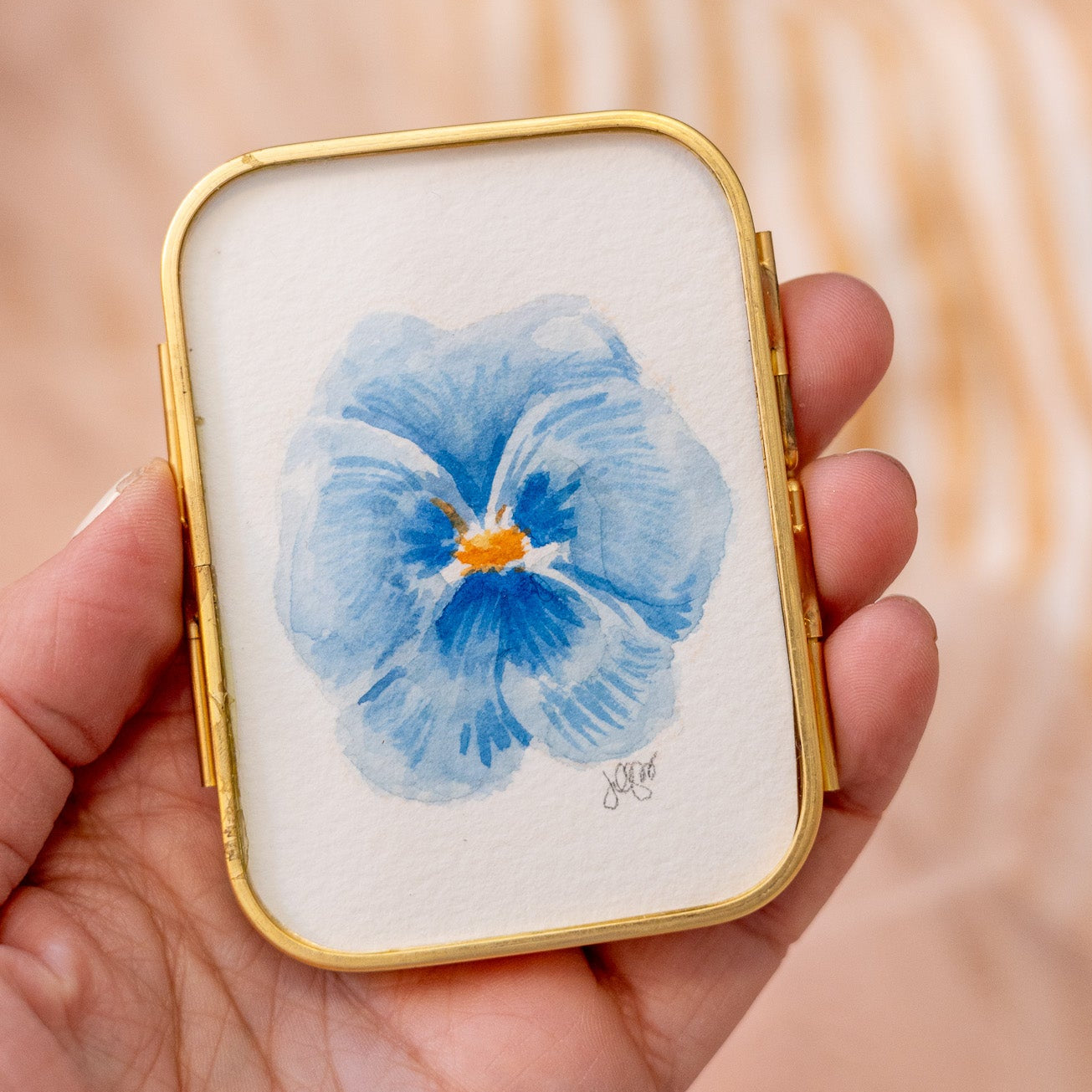 Small rectangular gold frame with a blue pansy flower painting held in a hand against a blurred background